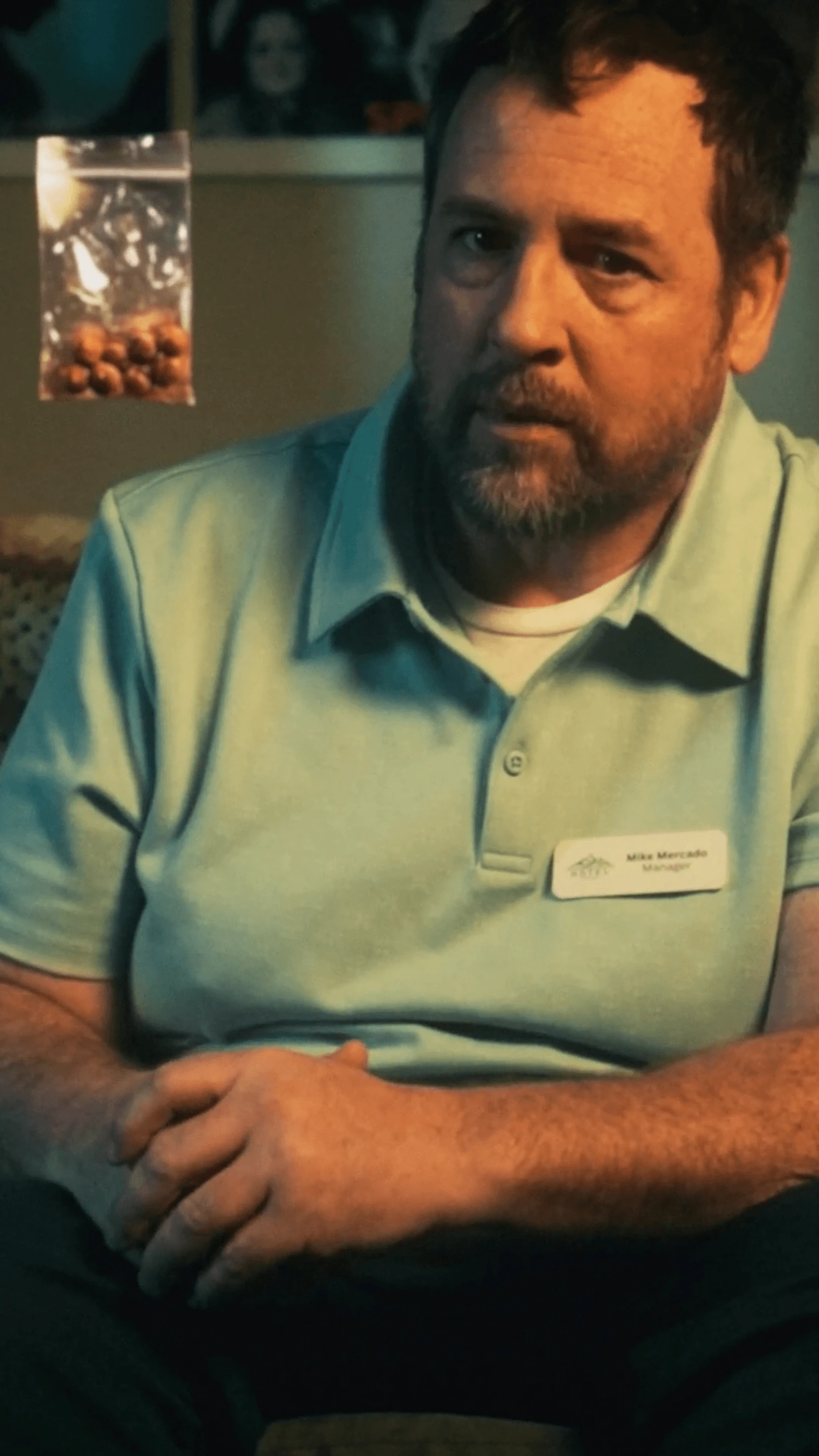 A man in a work shirt sits in a living room next to a bag of pills